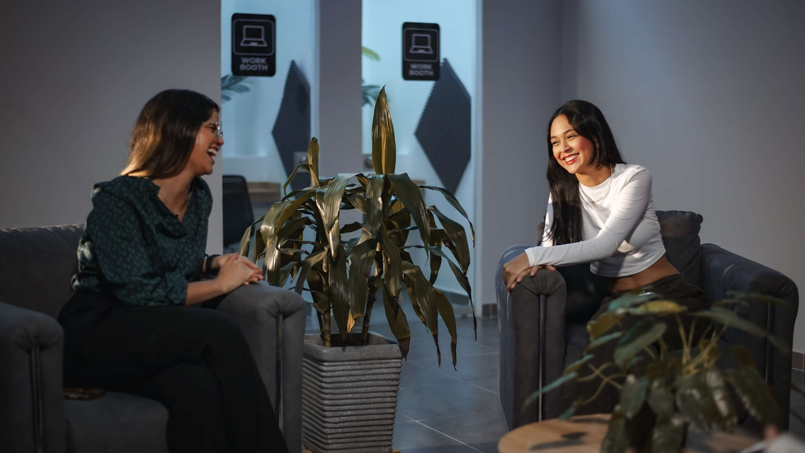 Two women sitting across from each other in a modern coworking lounge, laughing and enjoying a friendly conversation. The setting features comfortable gray chairs, indoor plants, and “work booth” signs in the background, conveying a relaxed yet professional atmosphere.
