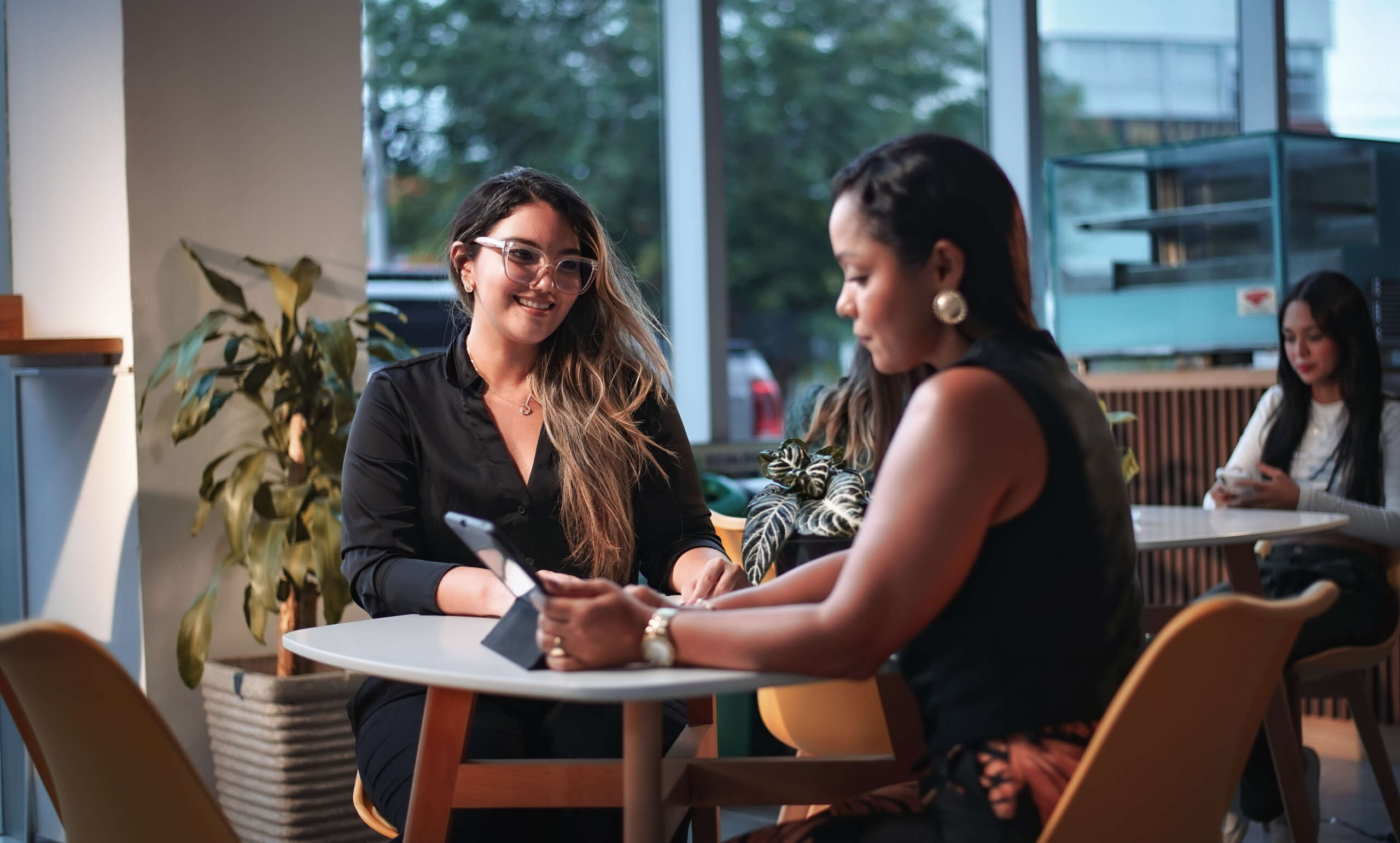 Two women sit at a table in a modern coworking café, engaged in a friendly conversation while using a tablet. Natural light streams through large windows behind them, with plants and other people working in the background, creating a warm and professional atmosphere.