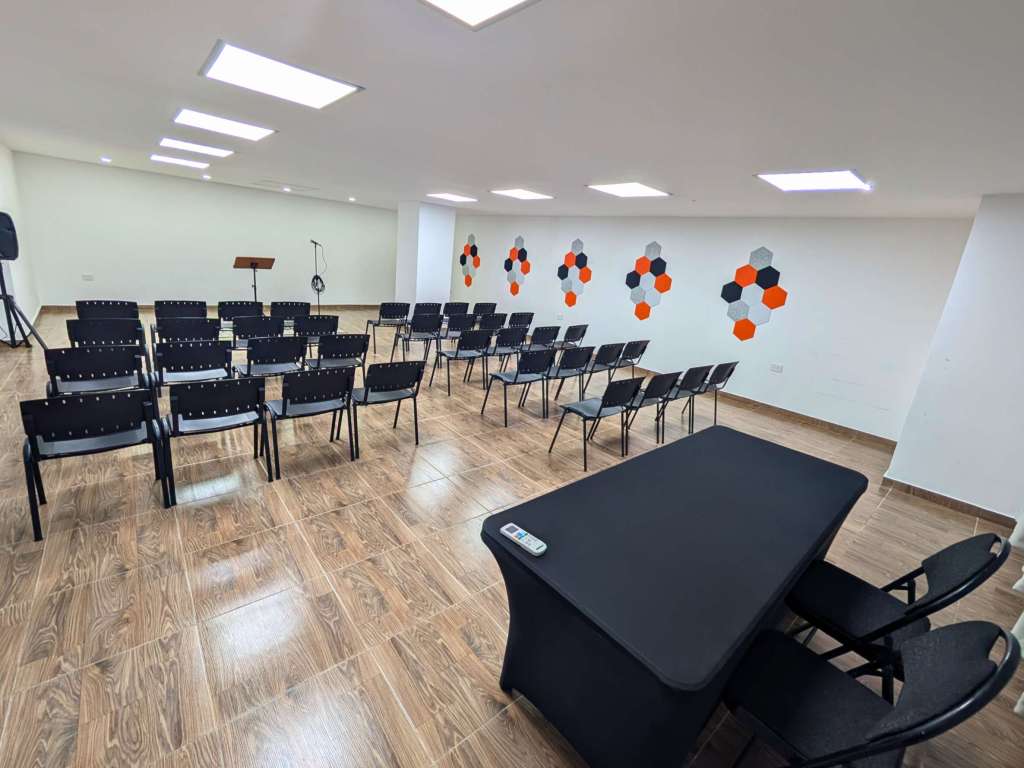 Espacio de conferencia en Coventus con sillas organizadas en filas, un podio con micrófono al frente y una mesa con mantel negro en primer plano. Las paredes blancas están decoradas con paneles hexagonales en tonos naranja, gris y negro. La sala cuenta con iluminación LED en el techo y un parlante en la esquina para sonido amplificado.