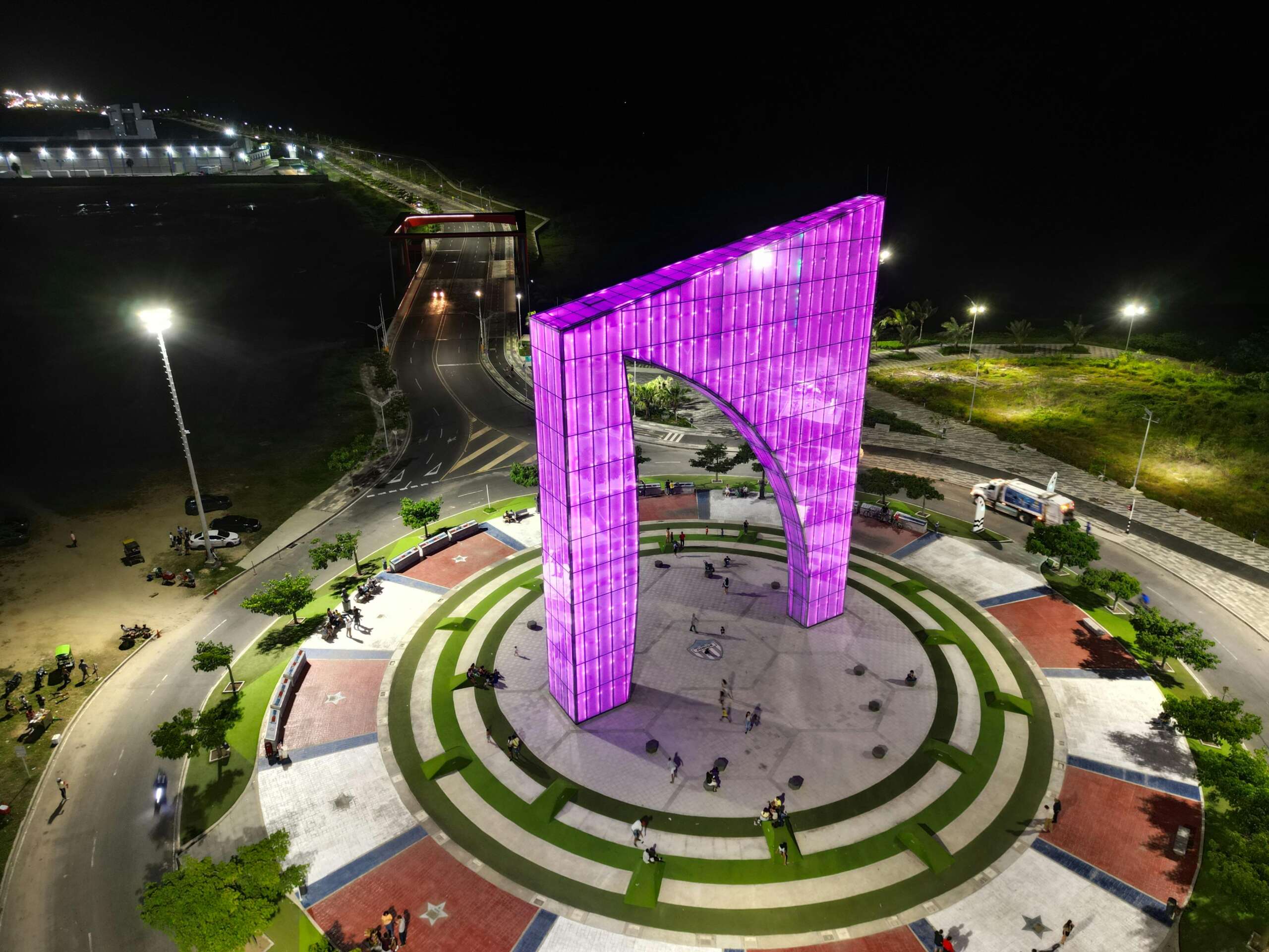 Aerial night view of La Ventana al Mundo monument in Barranquilla, Colombia, illuminated in bright purple lights. The large arch-shaped structure stands at the center of a circular plaza surrounded by walkways, greenery, and streetlights, symbolizing modernity and pride on the Caribbean coast.